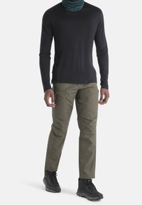 Mann trägt ein schwarzes Langarmshirt, einen dunkelgrünen Halswärmer, olivgrüne Cargo-Hosen und schwarze Wanderschuhe und steht vor einem weißen Hintergrund.