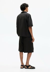 Camicia nera di cotone a maniche corte e pantaloni coordinati, entrambi con una vestibilità ampia. Abbinati a sandali neri con cinturini.