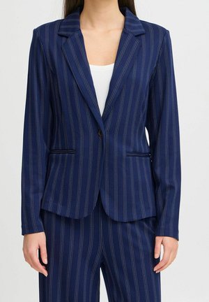 Kvinde iført marineblå blazerkjole med nålestriber og matchende bukser over hvid top, stående mod en ensartet lys baggrund.