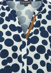 Blusa a maniche corte in tessuto leggero con pois blu navy su sfondo bianco. Presenta un collo a V e un accento in velluto marrone.
