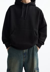 Svart huvtröja med framficka och snörning. Den har en avslappnad passform och är kombinerad med blå jeans.