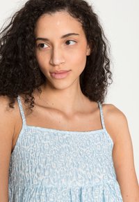 Vestido azul claro com alças ajustáveis, apresentando um padrão de tecido texturizado e corpete franzido. O design é fluido e descontraído.