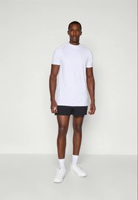 Mann steht frontal mit einem weißen Sport-T-Shirt, schwarzen Shorts, weißen Socken und weißen Sneakers vor einem schlichten Hintergrund.
