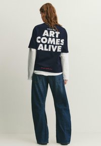 Person mit langen Haaren trägt ein marineblaues Hemd über weißen Langarmshirts, Jeans, mit dem Text „ART COMES ALIVE“ auf der Rückseite.
