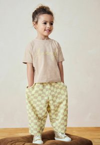 T-shirt beige à manches courtes avec "rire" en écriture jaune, associé à un pantalon ample à carreaux jaunes et marron et des baskets blanches.