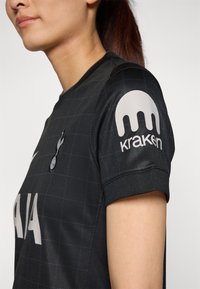 Maillot de sport noir à manches courtes, en tissu structuré, avec un motif en grille, arborant le logo "Kraken" en gris clair sur la manche droite.