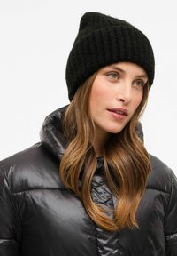 Beanie en tricot noir avec une texture côtelée, présente une pointe effilée. L'utilisateur porte une doudoune noire brillante avec une capuche, révélant des cheveux bruns doux.