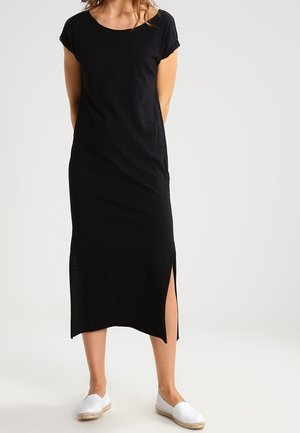 Frau trägt ein schwarzes Midikleid mit kurzen Ärmeln und einem seitlichen Schlitz, kombiniert mit weißen Espadrille-Schuhen zum Hineinschlüpfen, steht vor einem einfarbigen Hintergrund.