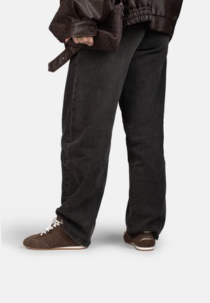 Persona che indossa jeans neri larghi, sneakers marroni con lacci bianchi e una giacca di pelle marrone scuro, vista dalla metà coscia in giù.