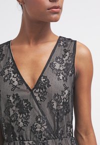 Robe en dentelle noire avec motifs floraux, un décolleté en V profond et une superposition texturée. Le tissu est transparent avec des détails subtils.