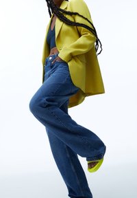 Femme aux longues tresses portant un blazer jaune, un crop top sombre, un jean bleu ample et des sandales jaune néon, posant devant un fond blanc.