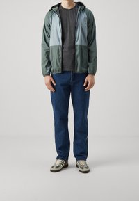 Veste légère à capuche en panneaux vert foncé et bleu clair, associée à un t-shirt gris et un jean bleu, complétée par des baskets vertes et marron.