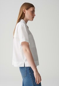 Weißes kurzärmliges Hemd mit lockerer Passform, mit Kragen, horizontalen Nahtdetails und seitlichen Schlitzen. Kombiniert mit blauen Jeans.