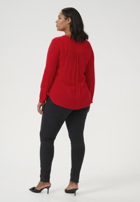 Camisa roja de manga larga con la parte posterior con pliegues, combinada con jeans ajustados negros y zapatos de tacón abiertos, vista desde atrás.