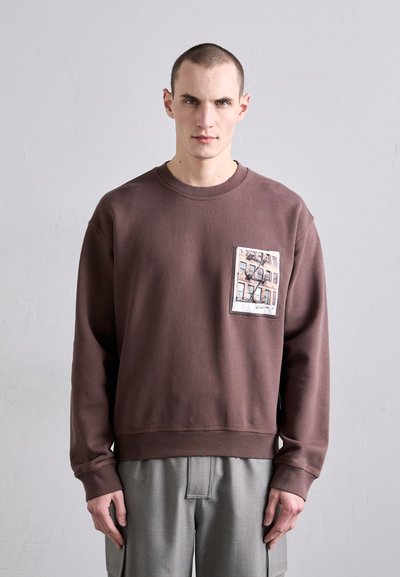 Sweatshirt marron avec un col rond, présente une étiquette rectangulaire avec une impression de bâtiment, texture douce et poignets et ourlet côtelés.