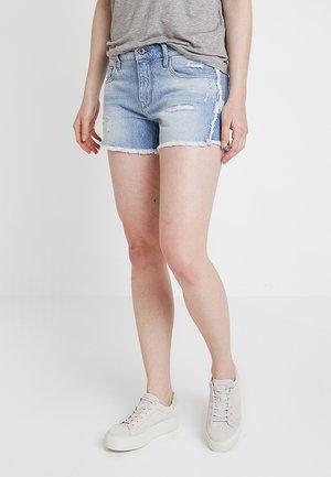 Person iført lyseblå slidte denimshorts, grå t-shirt og hvide platform sneakers står mod en ensfarvet hvid baggrund.