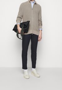 Beige stickad zip-up tröja, ljusblå kragdress under, mörka byxor och beige sneakers; håller en svart tote-väska med gröna remmar.