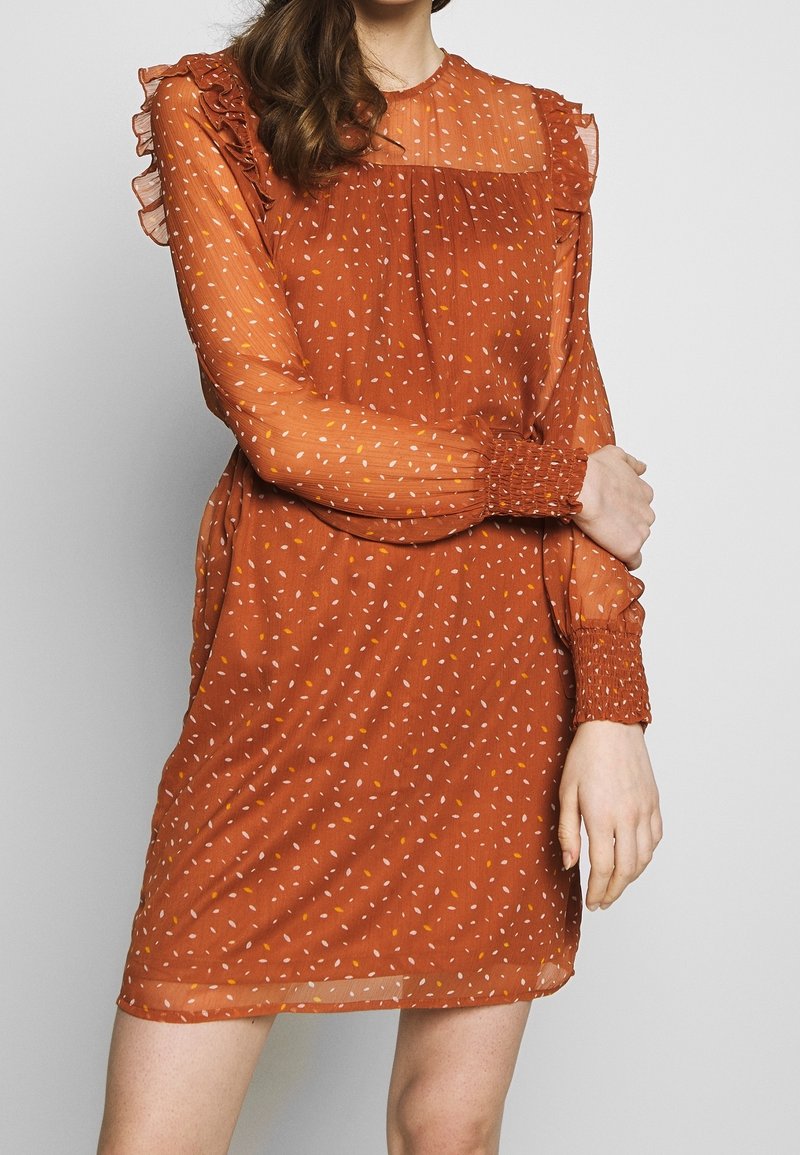 Robe à manches courtes de couleur rouille en tissu léger avec un col à volants et des poignets élastiqués, ornée de motifs tachetés blancs et jaunes.
