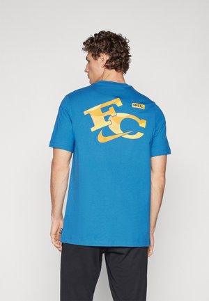 Blauwe katoenen T-shirt met gele graphics op de achterkant, waaronder "F.C." en het Nike-logo. Korte mouwen, relaxte pasvorm, soepele textuur.