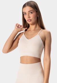 Sostén deportivo beige con tirantes ajustables, un escote en V, detalles fruncidos en el centro y una banda con logo en la parte inferior. Tela suave y elástica.