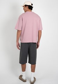 Homme vu de dos portant un t-shirt rose oversize, un short gris rayé aux genoux, des chaussettes blanches, des sandales beiges et une casquette de baseball blanche.