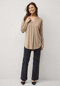 Beige langærmet top med V-halsudskæring, der har en afslappet pasform og rund kant; kombineret med mørke jeans og spidse sorte sko.