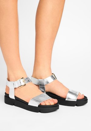 Srebrne metaliczne sandały z grubą czarną podeszwą, wyposażone w regulowane paski wokół kostki i wytłaczane logo na cholewce.