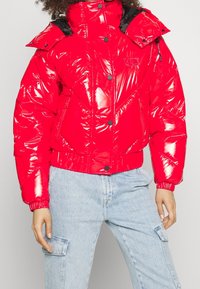 Veste puffer rouge avec une finition brillante, dotée d'un col haut, d'accents noirs, de boutons-pression et d'un motif matelassé en forme de losange.
