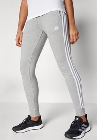 Sivé atletické legíny majú priliehavý strih, biele trojpruhy na bokoch a logo Adidas pri boku. V kombinácii s čiernymi športovými topánkami.