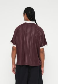 Camisa de manga curta numa tonalidade granada com padrão vertical, colarinho branco em contraste e um corte solto. Tecido macio e texturizado com uma marcação subtil.