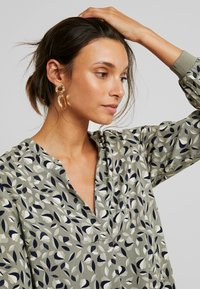 Blusa floral em verde, com decote em V e mangas longas. O padrão inclui formas em azul-marinho e creme, com detalhes texturizados. Brincos circulares em ouro.