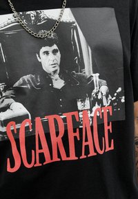 Sort bomulds T-shirt med et grafisk print af en sort-hvid scene fra "Scarface" med rød tekst nedenfor, der siger "SCARFACE." Detalje med kædenecklace.
