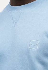 Άνδρας φοράει γαλάζιο φούτερ με στρογγυλή λαιμόκοψη και μικρό τετράγωνο λογότυπο «BOSS» στο στήθος.