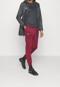 Person med svart Nike-jacka, svart t-shirt, vinröda Nike F.C. joggingsbyxor och svarta Nike-sneakers som står mot en enkel vit bakgrund.