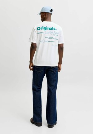 Homme portant une casquette bleu clair, un t-shirt blanc avec l'inscription « Originals », un jean bleu foncé et des chaussures marron, debout de dos sur un fond uni.