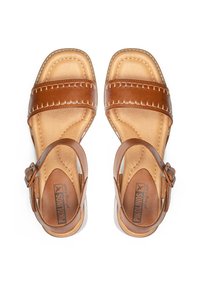 Sandalias de cuero marrón con dos correas anchas, bordes cosidos y una hebilla ajustable en el tobillo. Plantilla de cuero natural y suela translúcida.