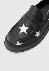 Svarta läderloafers med utskurna vita stjärnmönster, med ett band över vristen och en tjock gummisula för bra grepp.