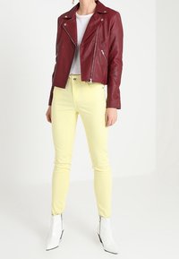 Giacca da biker in pelle borgogna con dettagli zip, indossata sopra una maglietta bianca e jeans skinny gialli. Completata con stivaletti bianchi alla caviglia.