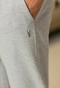 Graue Sweatpants aus weichem Material mit seitlicher Tasche und einem kleinen braunen Hirsch-Emblem, das auf dem Oberschenkel gestickt ist.