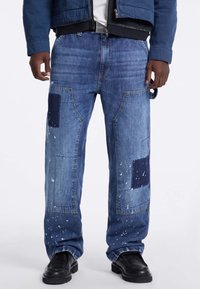 Jeans in denim con un design a patchwork con pezzi di stoffa blu scuro e blu chiaro, accenti di schizzi di vernice e una vestibilità rilassata con ampie tasche laterali.