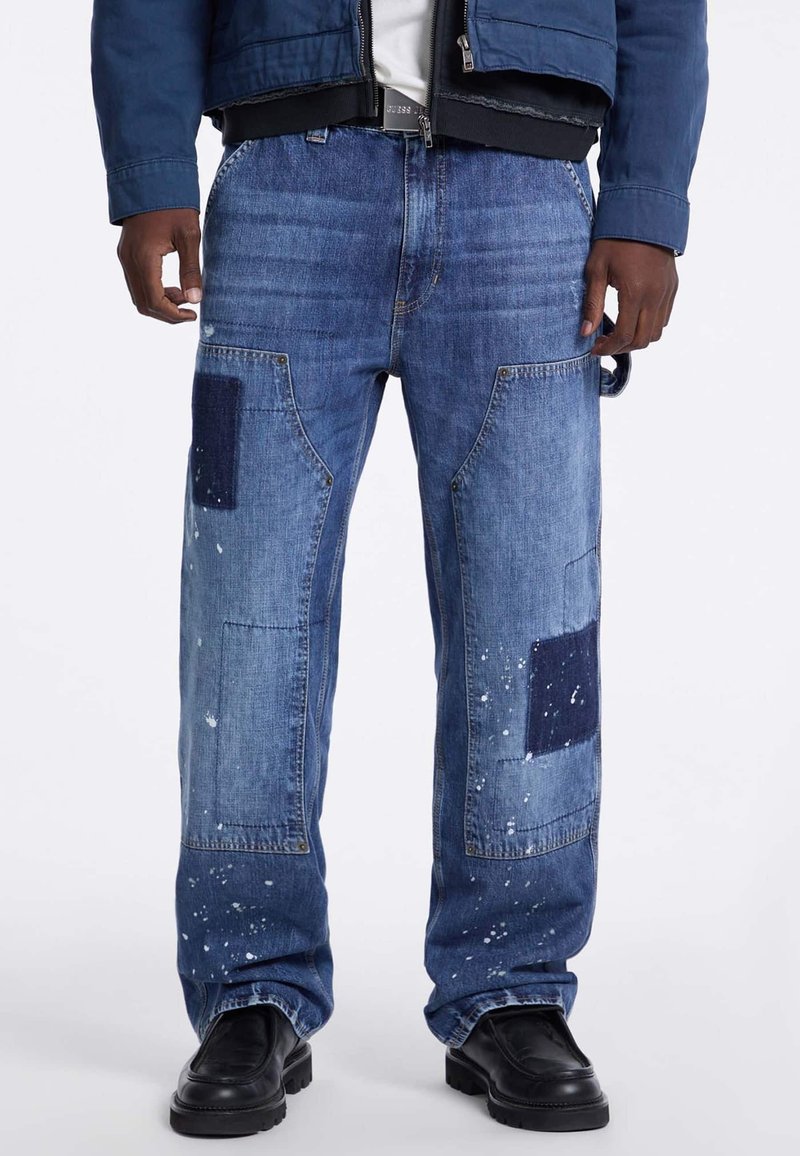 Jeans in denim con un design a patchwork con pezzi di stoffa blu scuro e blu chiaro, accenti di schizzi di vernice e una vestibilità rilassata con ampie tasche laterali.