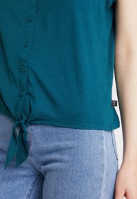 Teal kortärmad topp med knäppning framtill och knuten nederkant; ihop med ljusblå denimjeans med klassisk passform.