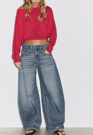 Femme portant un crop top rouge à épaules dénudées, manches longues, un jean bleu délavé à jambes larges et des sandales noires ouvertes à l'avant, debout sur un fond blanc.