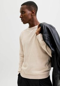 Maglione beige a maglia con scollo tondo e polsini a coste, abbinato a una giacca di pelle nera appoggiata sulla spalla.