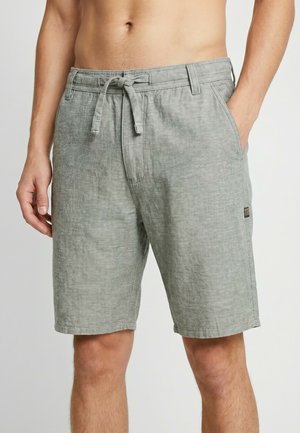 Man wearing light gray drawstring linen shorts with hand in pocket, standing against a plain light background.