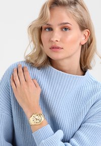 Guldring på håndleddet med rund urskive, tre dials og metalarmbånd; påført med en lys blå tekstureret striktrøje.