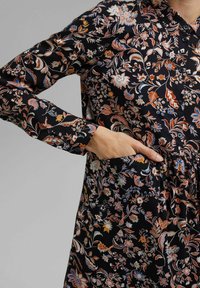 Blommig klänning i svart tyg med intrikata mönster i orange, blått och vitt. Har krage och sidofickor, långa ärmar.