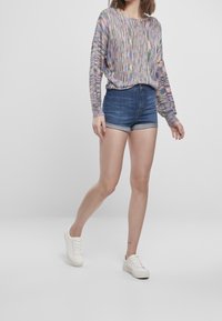 Donna che indossa un maglione a maniche lunghe multicolore, pantaloni shorts in denim blu arrotolati e sneakers bianche, che cammina su uno sfondo grigio chiaro uniforme.