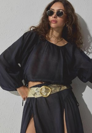 Zwarte doorschijnende blouse met opgeblazen mouwen, gecombineerd met een gouden metallic riem met een decoratieve gesp. Model draagt ronde zonnebril.