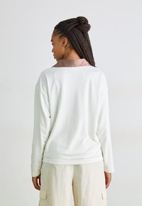 Weißes Langarmshirt aus Netzstoff mit einem kontrastierenden pinken Satin-Kragen, relaxed Fit und nahtlosen Bündchen. Getragen mit hellbeigen Hosen.